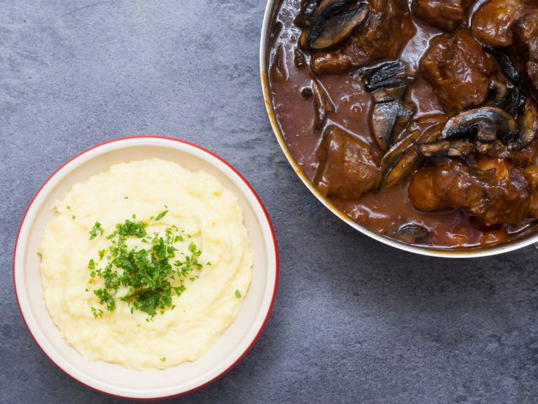 En skål med kartoffelmos toppet med hakket persille ved siden af en gryde med kødstykker og svampe i en mørk sovs på en grå bordplade.