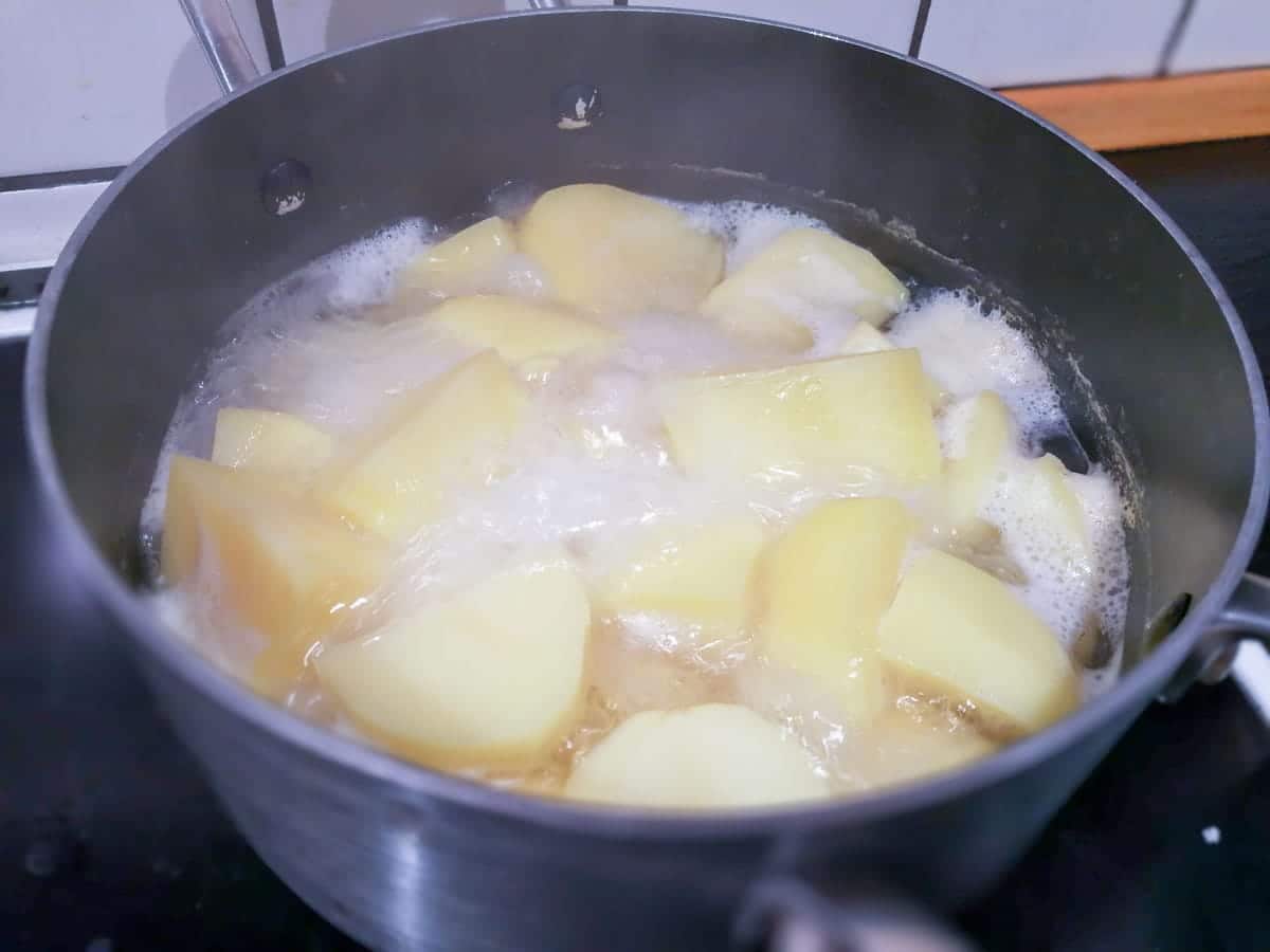 Kartofler koger i en stor gryde på et komfur i et køkken. Vandet bobler rundt om de delvist skrællede kartofler.