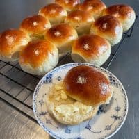 Gyldne burgerboller ligger på en rist; en åben bolle med smør på en blå-hvid tallerken står fremme. Alt befinder sig på en bordplade i metal.