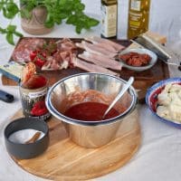 En metal skål med tomatsauce står på et træbræt med ingredienser som ost