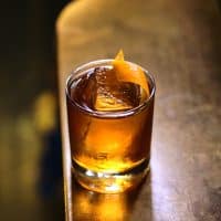 Et glas med en brun cocktail toppet med en stor isterning og et appelsinskal står på et træbord i et dæmpet oplyst rum.