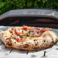 En pizza med sprød skorpe og skinke bages på en metalrist foran en moderne pizzaovn med "Ooni" skrevet på