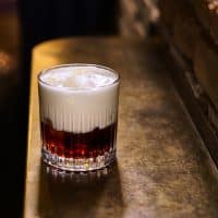 Et glas med en to-lags drink fyldt med isterninger står på en metaloverflade i et svagt oplyst rum ved en murstensvæg.
