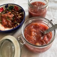 Krukker med rabarberkompot står på et bord. En skål med yoghurt og rabarberkompot er ved siden af. Bordet er dækket med en hvid dug.