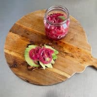 Et stykke rugbrød med avocado og syltede rødløg ligger på et træskærebræt